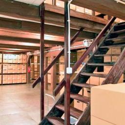 Archiwa Opolskie Sp. z o.o. - Magazyn z drewnianymi regałami i schodami, wypełniony kartonami z archiwum dokumentów, widoczne drewniane belki stropowe.