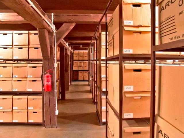 Korytarz archiwum z rzędami metalowych regałów wypełnionych kartonowymi pudłami, widoczny czerwony gaśnica umieszczona na drewnianym słupie.