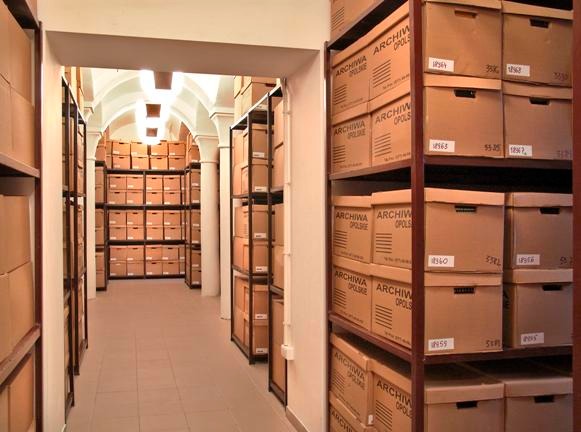 Korytarz archiwum z rzędami regałów wypełnionych kartonami z napisem 'Archiwa Opolskie', widok w głąb pomieszczenia z kolumnami i łukowym sklepieniem.