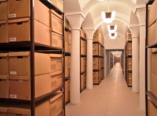 Długi korytarz archiwum z rzędami regałów wypełnionych kartonami oznaczonymi napisem 'Archiwa Polskie', oświetlony lampami i wsparty kolumnami.