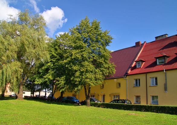 Żółty budynek z czerwoną dachówką widziany z perspektywy trawnika z drzewami, samochody zaparkowane wzdłuż budynku.