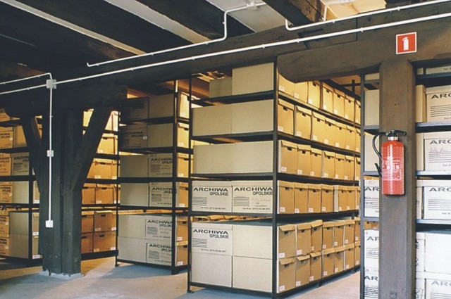 Regały magazynowe wypełnione kartonami z napisem 'Archiwa Opolskie' w pomieszczeniu z drewnianymi belkami stropowymi i instalacją elektryczną pod sufitem, dodatkowo widoczna gaśnica.