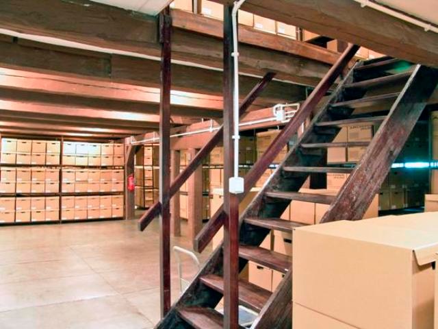 Magazyn z drewnianymi regałami i schodami, wypełniony kartonami z archiwum dokumentów, widoczne drewniane belki stropowe.