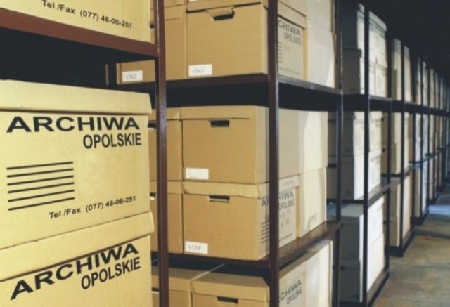 Rzędy metalowych regałów wypełnione kartonowymi pudłami archiwizacyjnymi, oznaczonymi etykietami i logo Archiwa Opolskie, w składnicy akt.