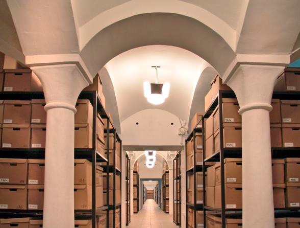 Długi korytarz piwniczny z rzędami regałów wypełnionych kartonowymi pudłami, oświetlony lampami w stylu art deco, z kolumnami podtrzymującymi sklepienie łukowe.