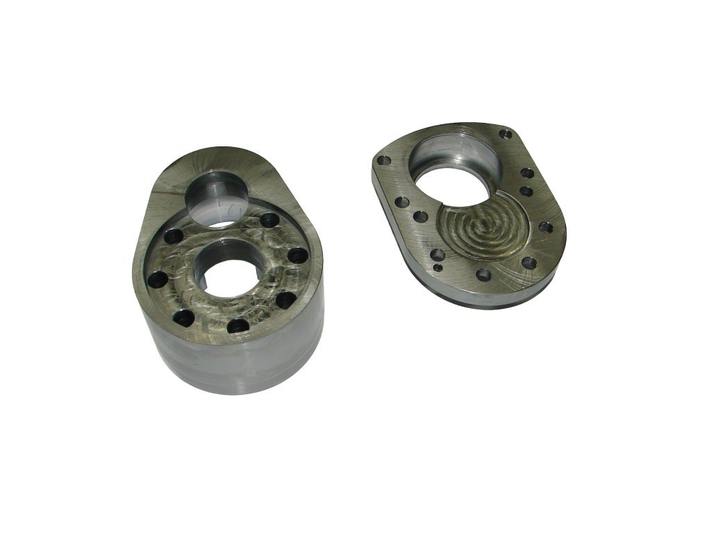 Dwie metalowe obudowy z otworami, jedna z spiralnym wzorem wewnątrz otworu, widoczne ślady obróbki.