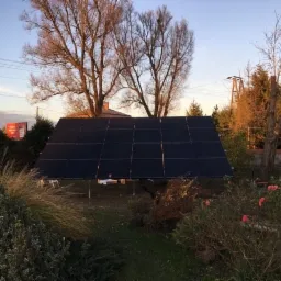 Czarny panel fotowoltaiczny na metalowej konstrukcji, ustawiony w ogrodzie z drzewami i krzewami o zachodzie słońca.