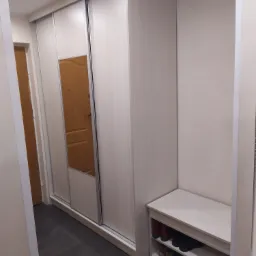 Jasna szafa wnękowa z przesuwnymi drzwiami, jednym lustrzanym, w przedpokoju z ciemną podłogą i siedziskiem na buty.