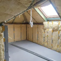 Poddasze w trakcie ocieplania wełną mineralną, widoczne profile metalowe, okno dachowe i fragment ściany z widocznymi śladami wcześniejszych prac.
