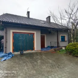 Przygotowania do malowania elewacji domu jednorodzinnego: okna i brama garażowa zabezpieczone folią ochronną, rozłożone płachty, widoczny wentylator osuszający.