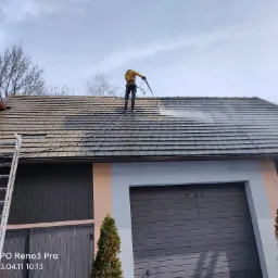 Pracownik w żółtej kurtce czyści dach z brudu za pomocą myjki ciśnieniowej, stojąc na nim w specjalnych butach z szeroką podeszwą. Dwie drabiny opierają się o ścianę budynku z szarą bramą garażową.