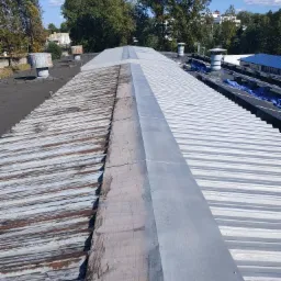 Porównanie dwóch połaci dachu: odnowiona, pomalowana na biało blacha trapezowa po prawej i stara, skorodowana blacha po lewej stronie, oddzielone szarym pasem. W tle drzewa i błękitne niebo.