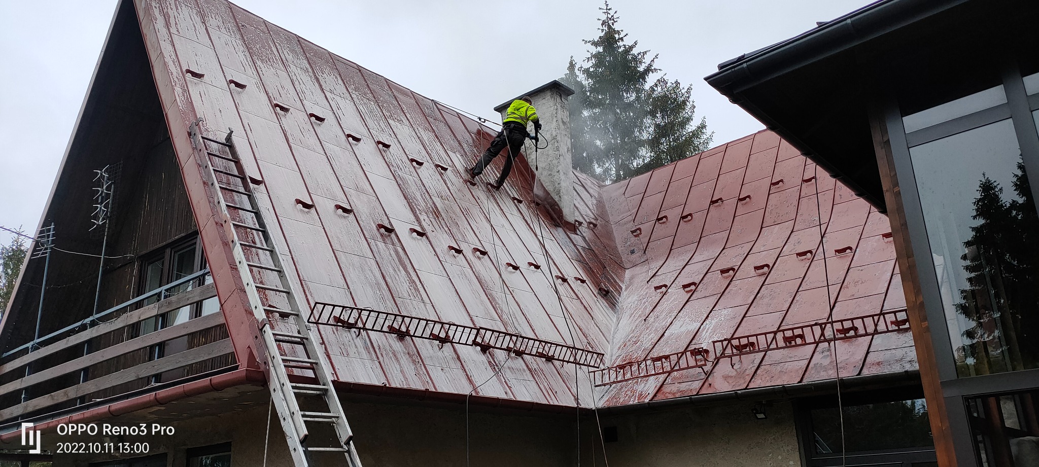 Pracownik w jaskrawej kamizelce czyści dach z metalowej blachy za pomocą myjki ciśnieniowej, widoczny komin, drabina oparta o dach, budynek z drewnianymi elementami w tle.