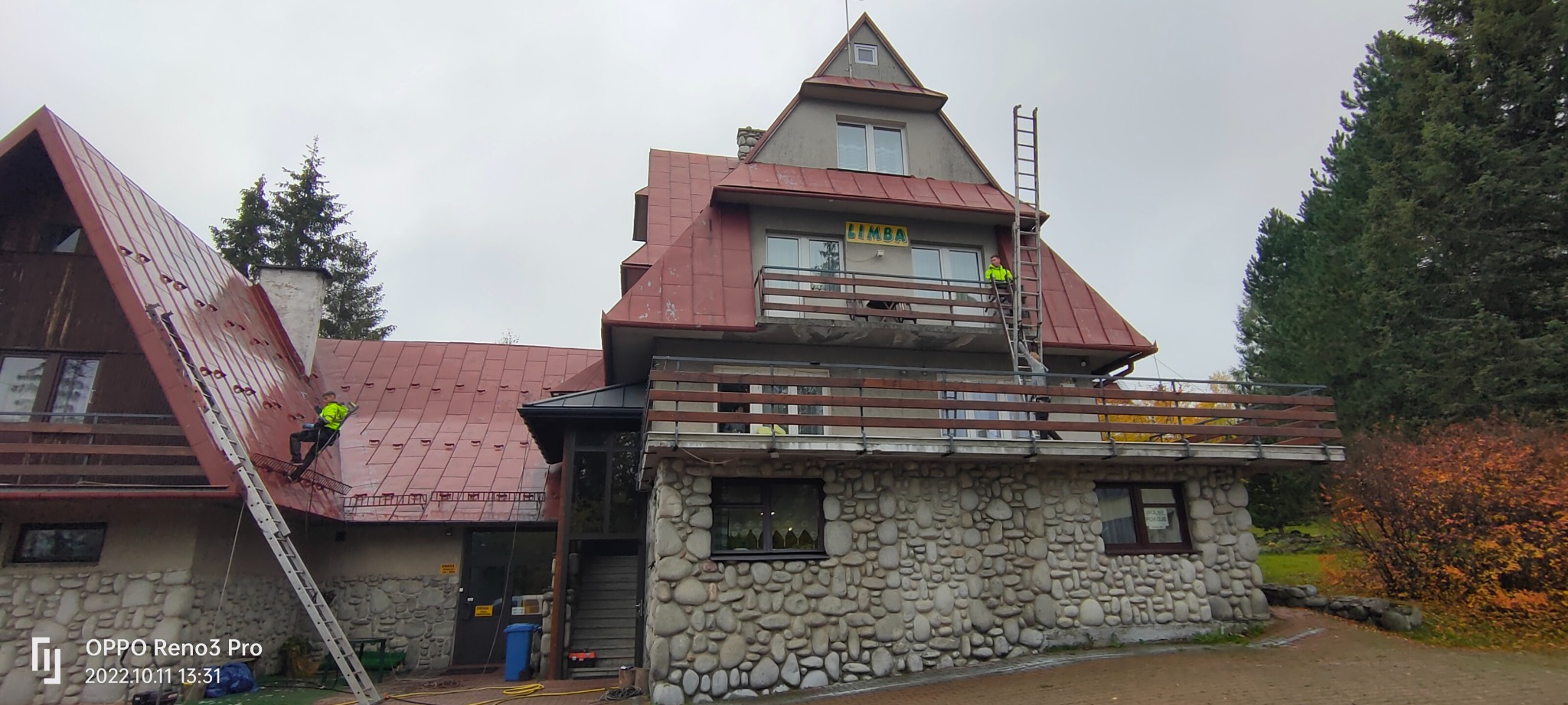 Pracownik w jaskrawej kamizelce na drabinie przy budynku z kamienną elewacją i czerwonym dachem, drugi pracownik na dachu z narzędziem w ręku.