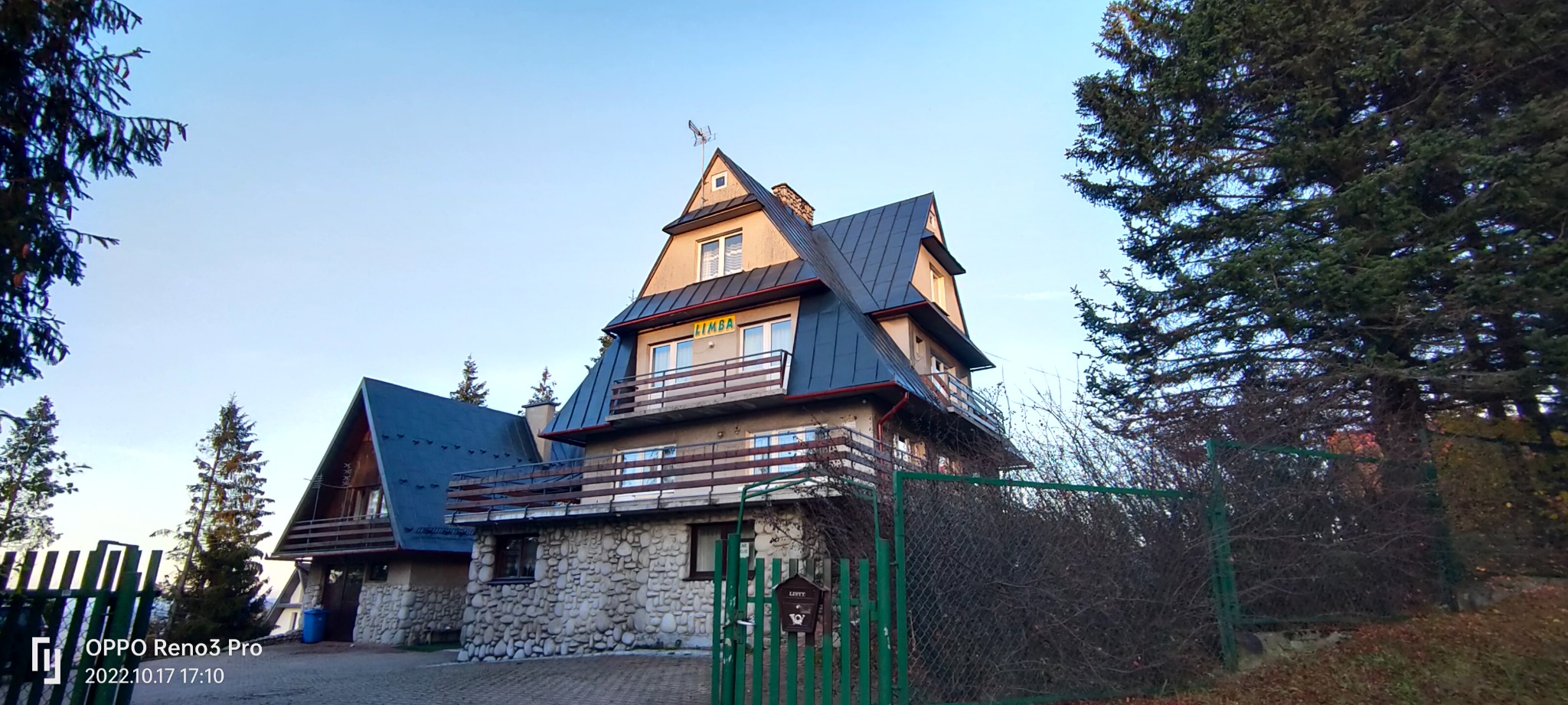 Dwupiętrowy budynek z kamienną podstawą i ciemnym dachem, widoczny zza zielonego ogrodzenia i drzew, z szyldem 'LIMBA' nad balkonem.