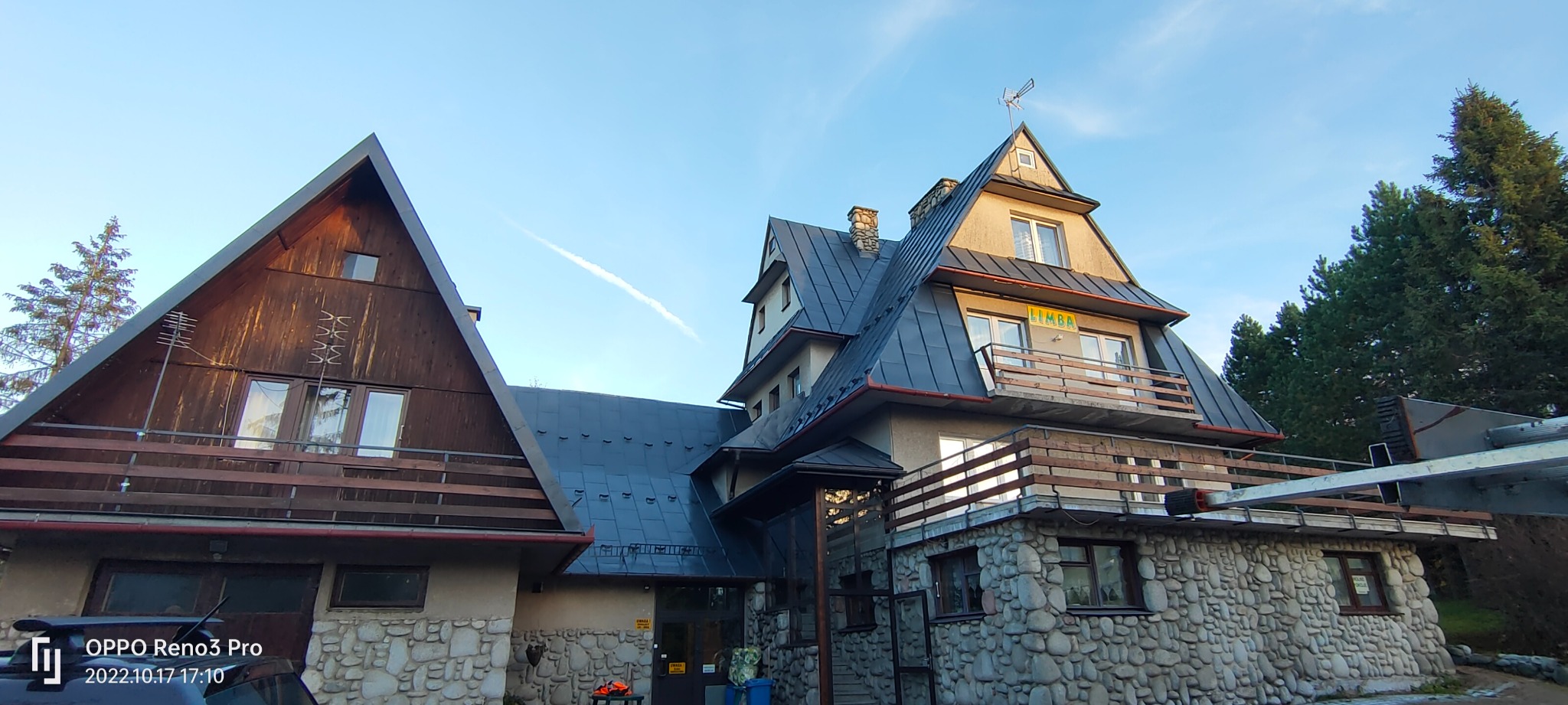 Wielopoziomowy budynek z ciemnym dachem blaszanym i kamienną elewacją, widoczny z dołu pod kątem, z fragmentem drabiny na pierwszym planie i smugą kondensacyjną na błękitnym niebie.