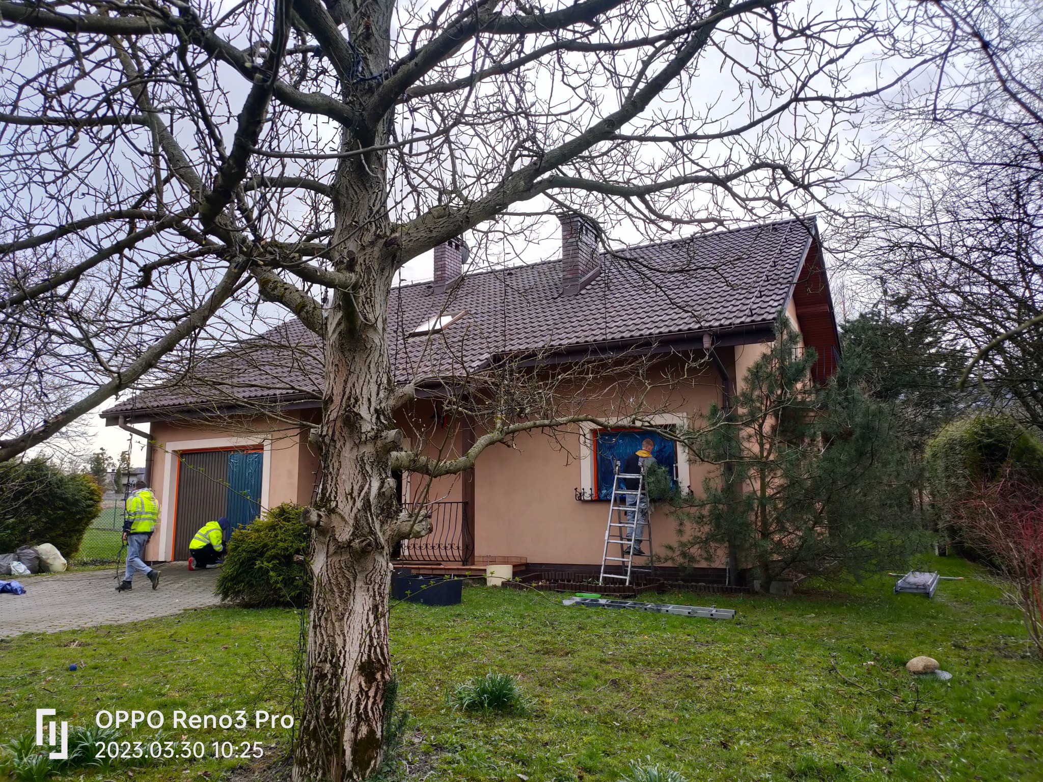 Pracownik na drabinie zabezpiecza okno folią przed rozpoczęciem prac na elewacji domu jednorodzinnego z brązowym dachem, widoczne narzędzia i drugi pracownik w odblaskowej kamizelce obok garażu.