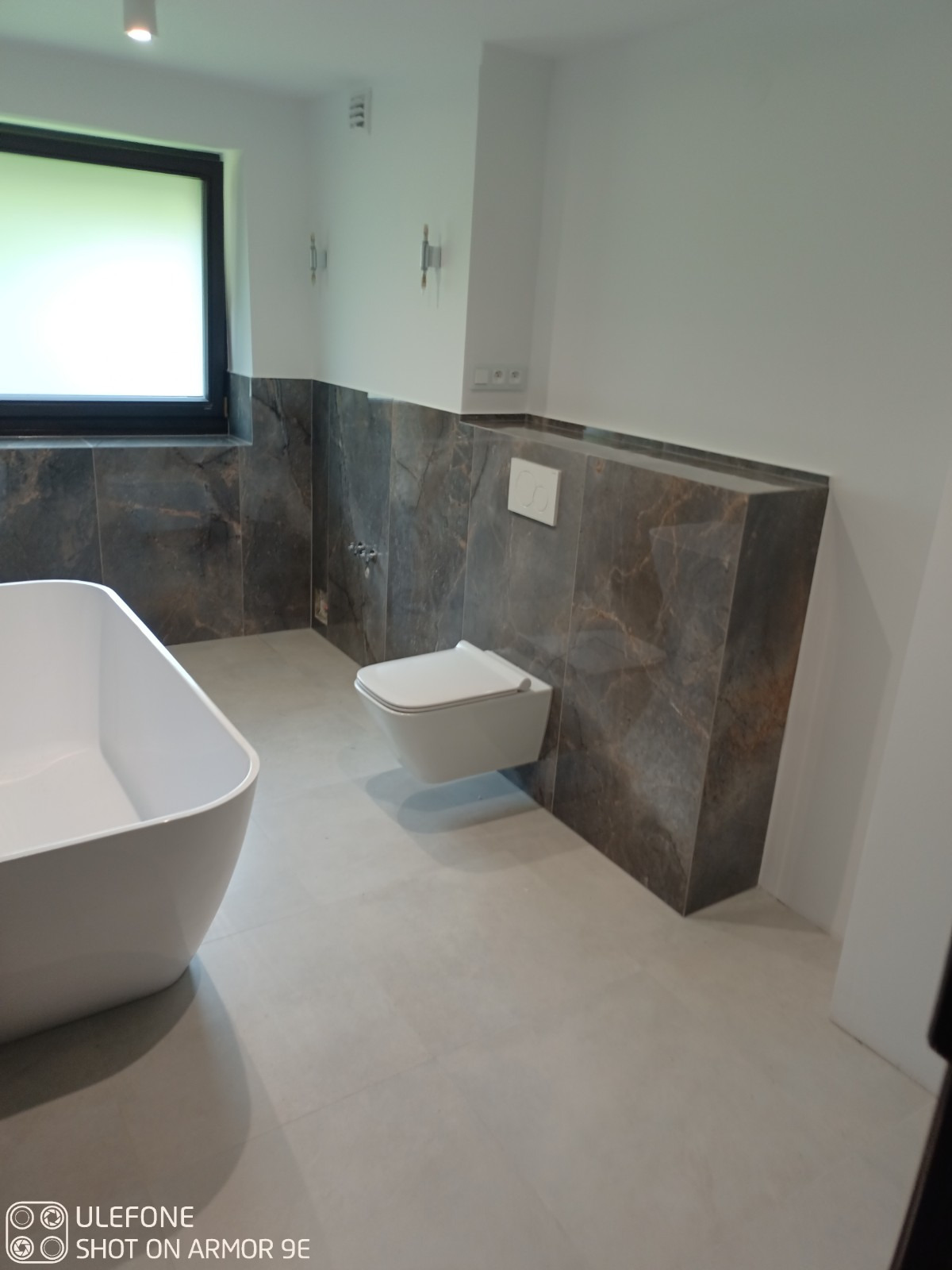 Nowoczesna łazienka z ciemnymi, marmurkowymi płytkami na ścianach do połowy wysokości, podwieszana toaleta, wolnostojąca wanna i jasna podłoga