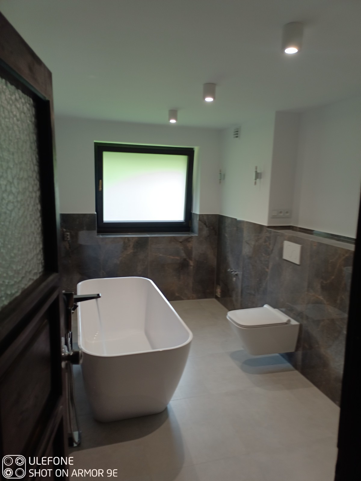 Nowoczesna łazienka z wolnostojącą wanną napełnianą wodą, podwieszaną toaletą i marmurowymi płytkami do połowy ściany, oświetlona punktowymi lampami sufitowymi. Widoczny fragment drewnianej ramy...