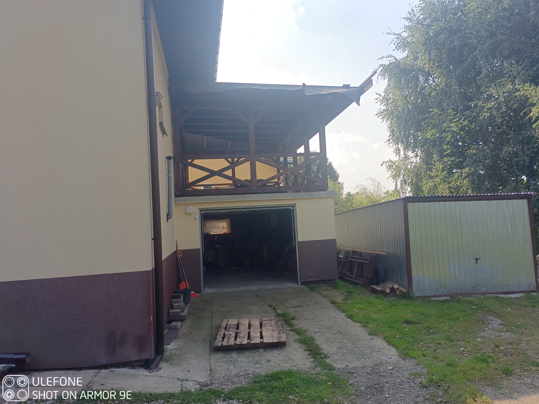 Konstrukcja drewnianej altany o powierzchni 38m2, widoczna z zewnątrz budynku, z garażem w tle i paletą na podjeździe.