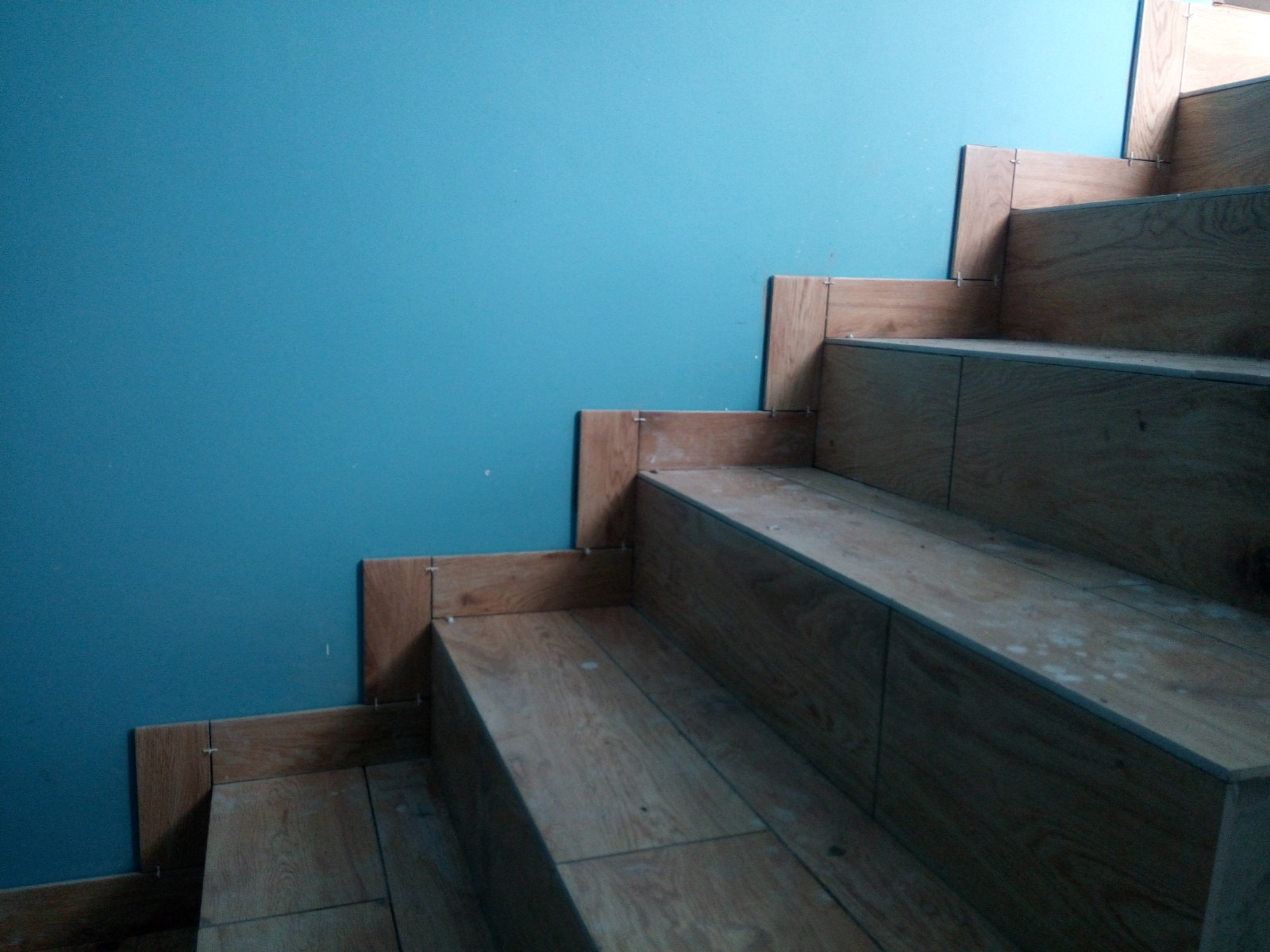 Kładzenie płytek imitujących drewno na schodach przy turkusowej ścianie, widoczne krzyżyki dystansowe.