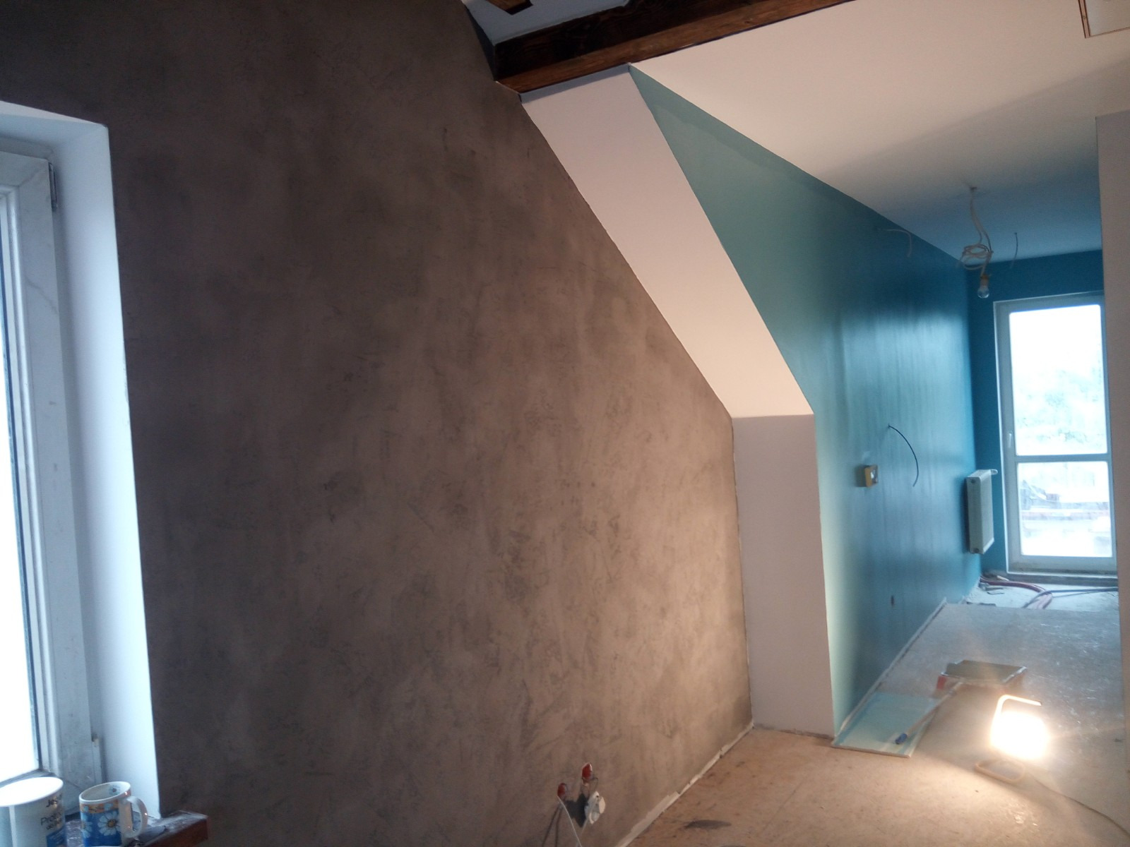 Pokój po częściowym wykończeniu: szara ściana z efektem dekoracyjnym obok białego okna, turkusowa ściana z grzejnikiem i wystającymi kablami, podłoga zabezpieczona folią, widoczne narzędzia i lampa...