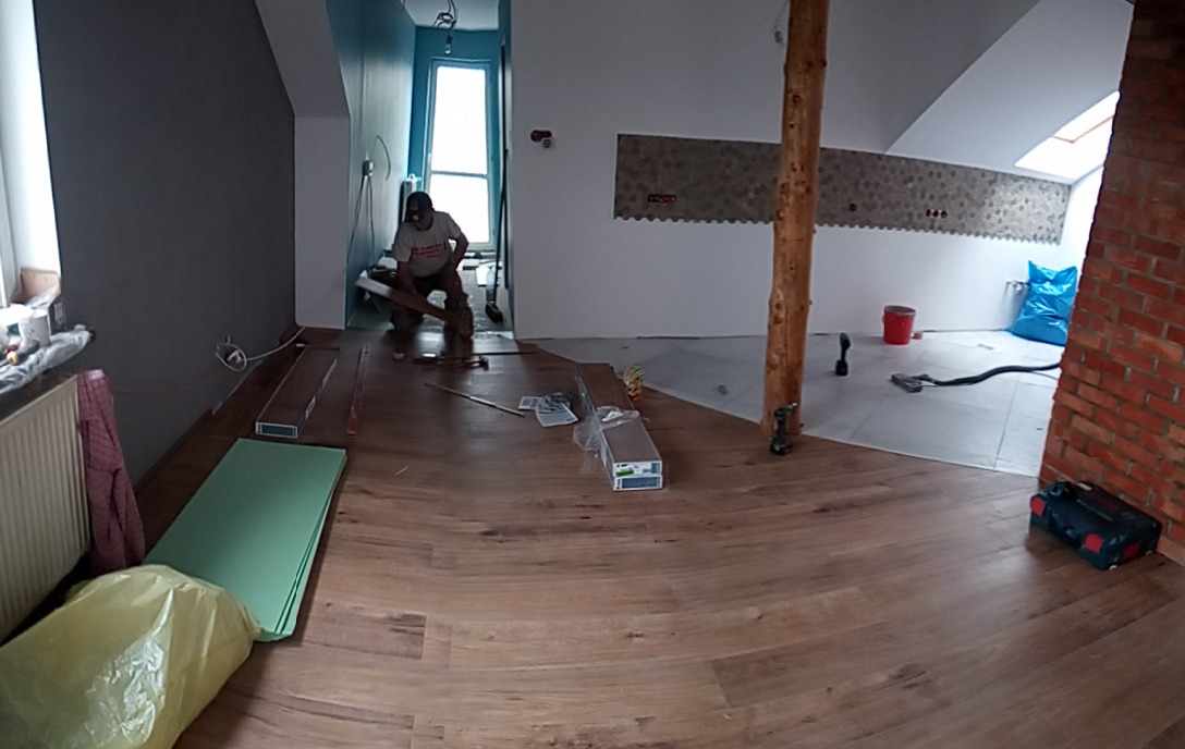 Układanie paneli podłogowych w jasnym wnętrzu z drewnianą kolumną i ceglanym narożnikiem; widoczny pracownik z narzędziami oraz materiały budowlane.