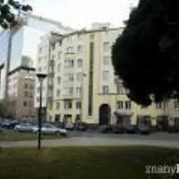 Widok na budynek mieszkalny z żółtą elewacją i łukowym wejściem, otoczony zielonym trawnikiem i drzewami, z nowoczesnym szklanym biurowcem w tle.