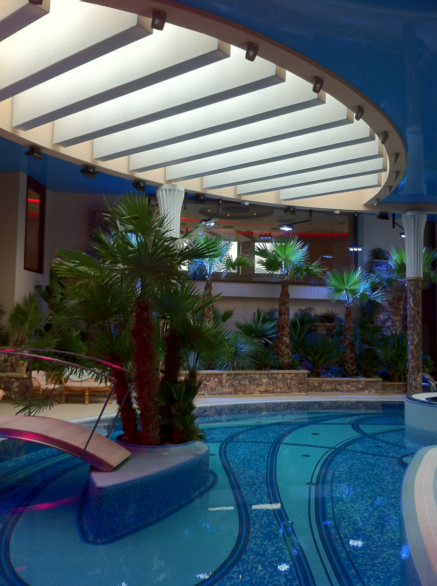 Kryty basen z mozaiką w odcieniach błękitu, z małym mostkiem i palmami, oświetlony dekoracyjnym sufitem z białymi panelami i niebieskim tłem.