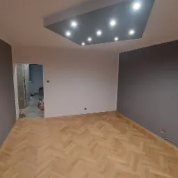 Pokój po remoncie w Zabrzu z parkietem w jodełkę, pomalowanymi na szaro ścianami i sufitem z oświetleniem punktowym. Widok na otwarte drzwi do kolejnego pomieszczenia w trakcie prac.