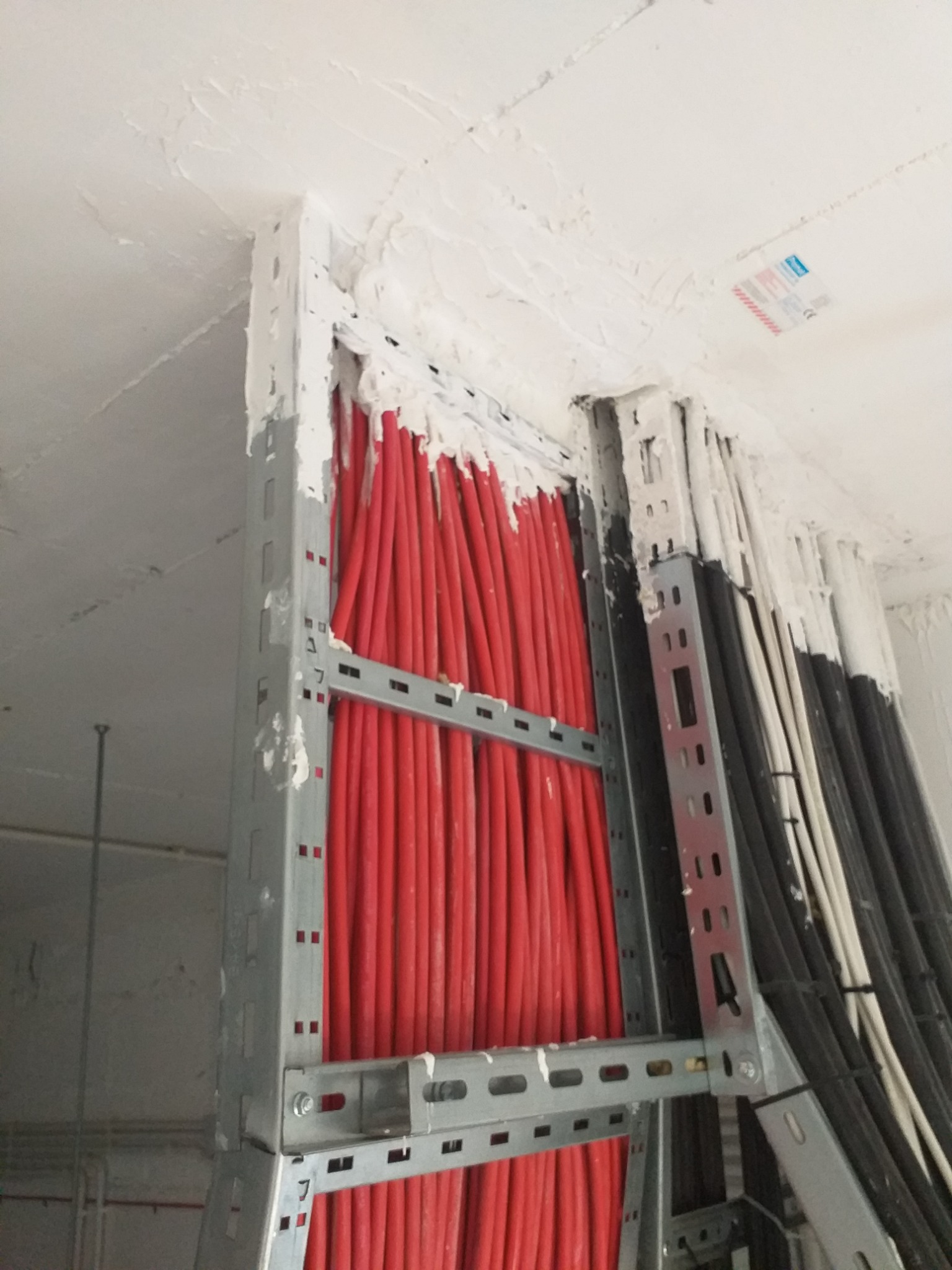 Pionowy układ czerwonych przewodów w metalowej konstrukcji, uszczelnionych gipsem przy suficie, obok wiązka czarnych i białych kabli.