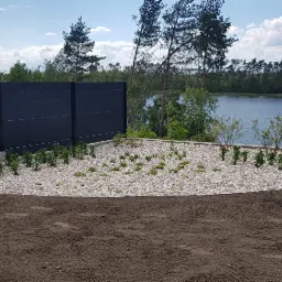 Nowo założony ogród z białymi kamieniami, młodymi roślinami i czarnym ogrodzeniem na tle jeziora i lasu.