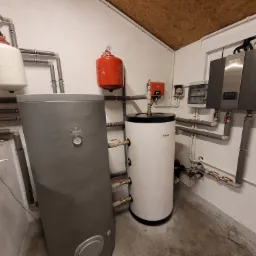 Instalacja w kotłowni z szarym i białym zbiornikiem akumulacyjnym, czerwonym naczyniem przeponowym, szarą jednostką pompy ciepła HEIKO i licznymi rurami z izolacją termiczną.