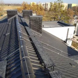 Czarny dach z zamontowaną instalacją odgromową, widoczne kominy z czarnej cegły i metalowe pomosty komunikacyjne, budynki w tle.