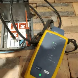 Tester kabli sieciowych Fluke DSX-600 z podłączonymi przewodami i otwartą puszką elektryczną na tle ceglanej ściany. Widoczny śrubokręt wewnątrz puszki.