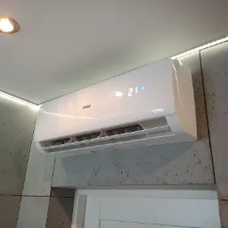 Biały klimatyzator Haier zamontowany na szarej ścianie z efektem betonu, z widocznym wyświetlaczem temperatury 21 stopni Celsjusza i oświetleniem LED.