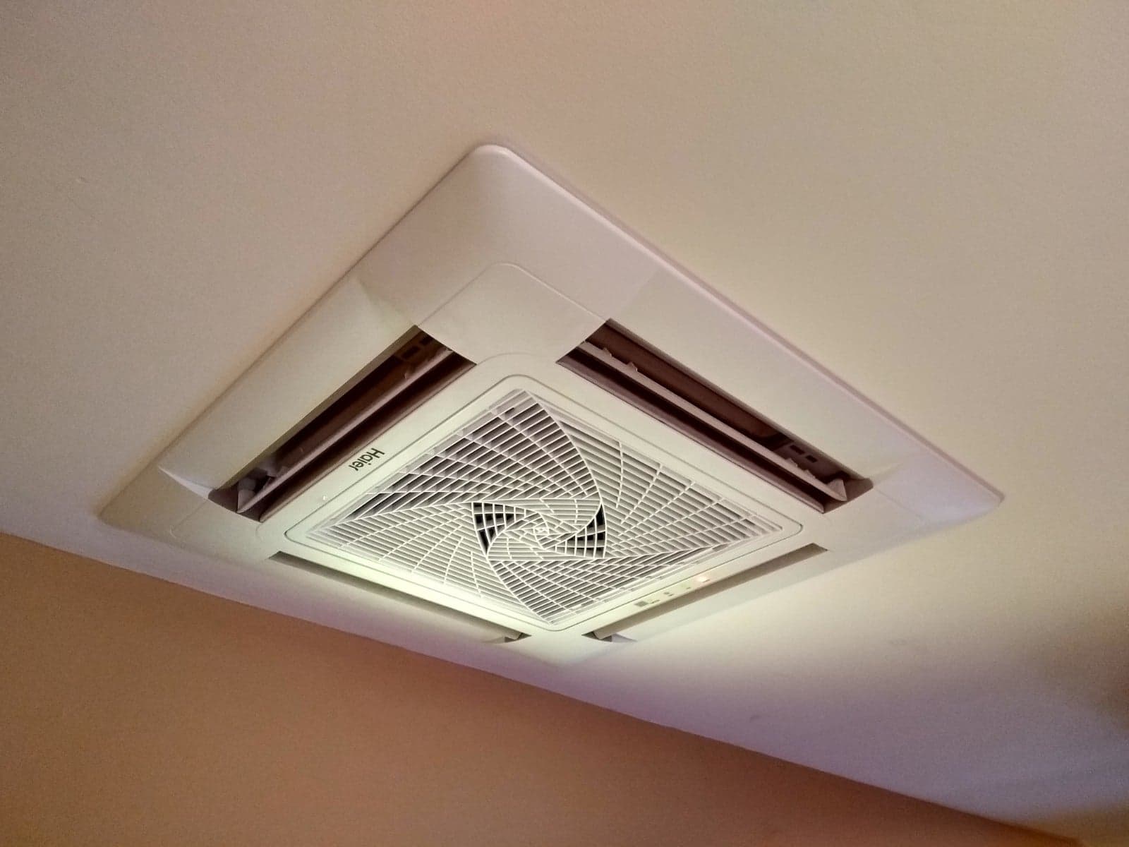 Kasetonowa jednostka klimatyzacji Haier zamontowana w suficie pomieszczenia, widok od dołu, z widocznymi kratkami wentylacyjnymi i logo producenta.