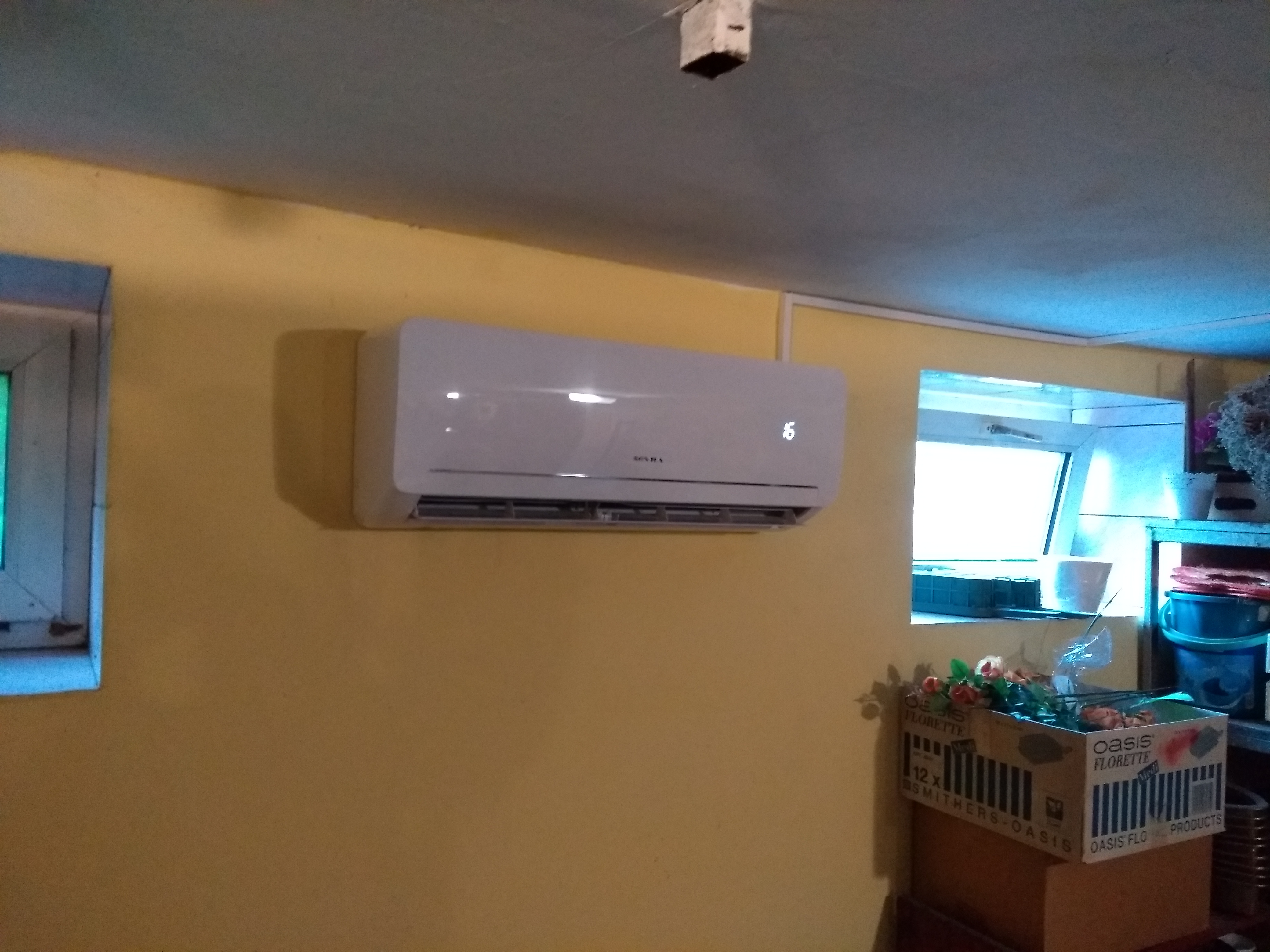 Biała jednostka klimatyzacji zamontowana na żółtej ścianie w pomieszczeniu z dwoma oknami, wyświetlająca temperaturę 16 stopni Celsjusza.