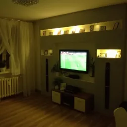 Salon z nowoczesną zabudową ściany telewizyjnej, podświetlane półki z dekoracjami, drewniana podłoga i grzejnik pod oknem z firankami.