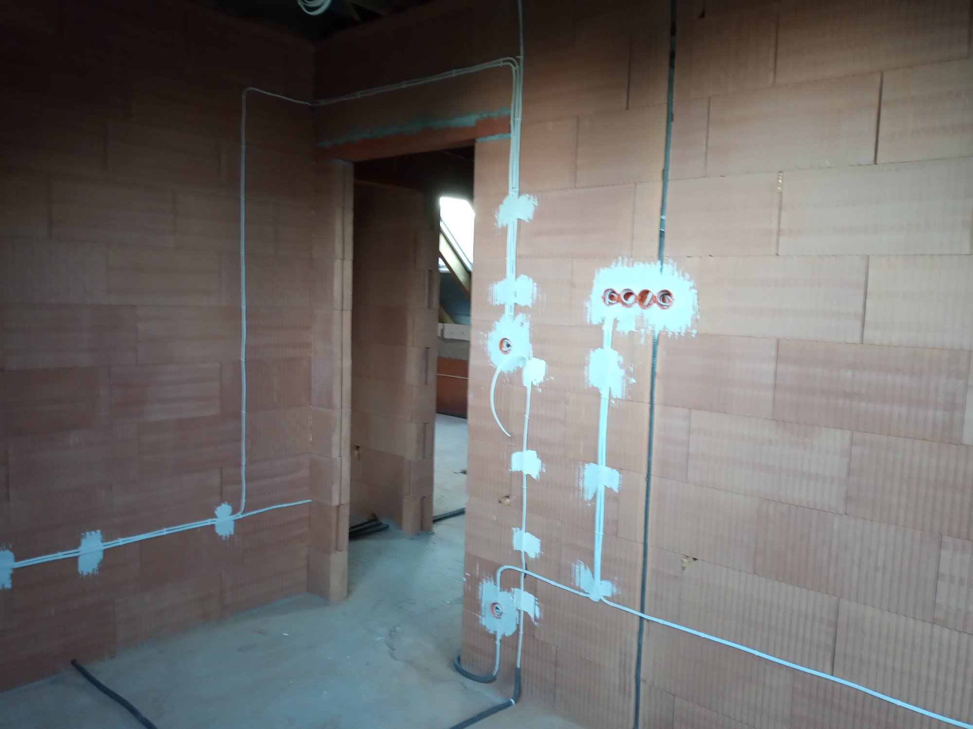 Instalacja elektryczna w surowym budynku z czerwonej cegły: białe kable poprowadzone po ścianach, puszki elektryczne częściowo zamaskowane gipsem.