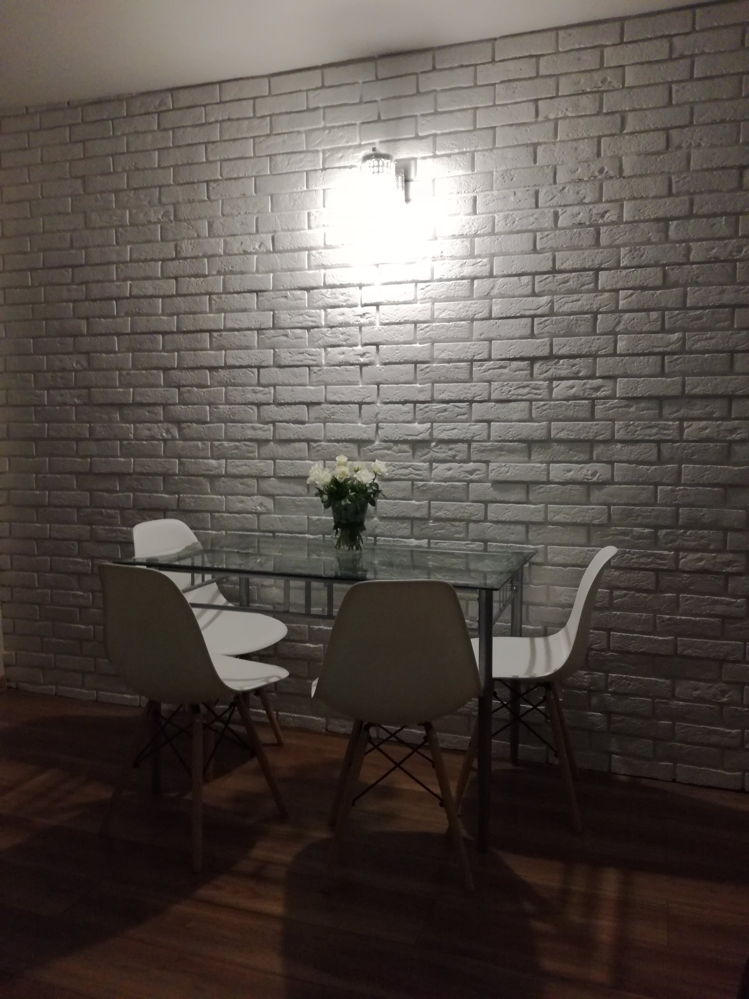 Jadalnia z białymi krzesłami, szklanym stołem i dekoracyjną ścianą z białej cegły podświetloną lampą.