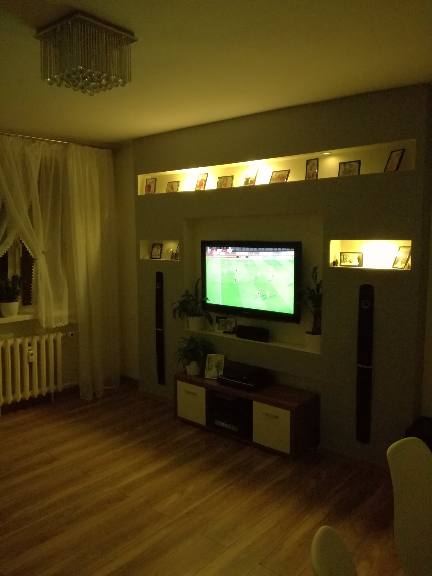 Salon z nowoczesną zabudową ściany telewizyjnej, podświetlane półki z dekoracjami, drewniana podłoga i grzejnik pod oknem z firankami.