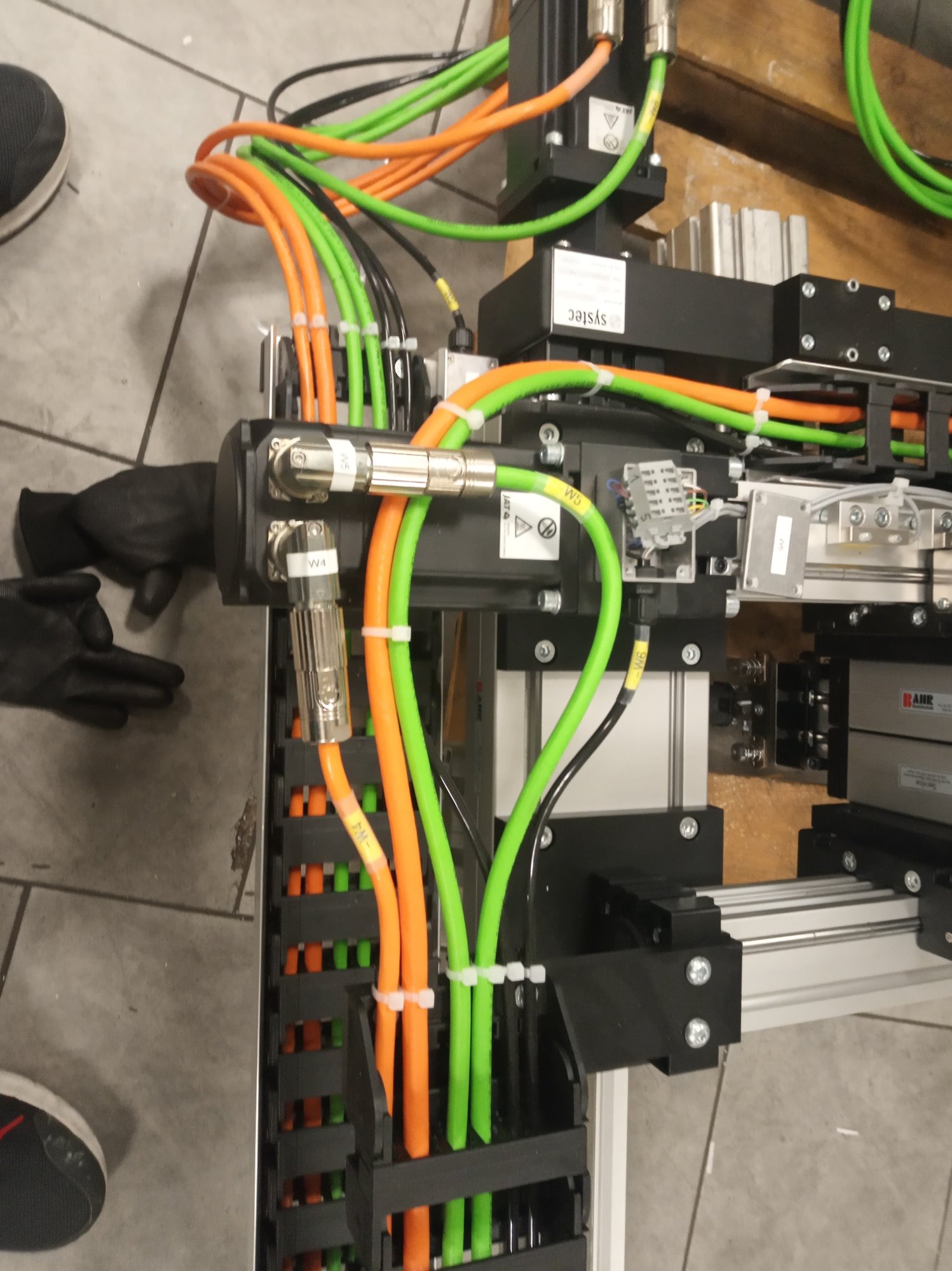 Szczegółowe ujęcie instalacji elektrycznej z licznymi przewodami w kolorach pomarańczowym i zielonym, uporządkowanymi w czarnych korytach kablowych, z widocznymi złączami i etykietami, obok dłoni...