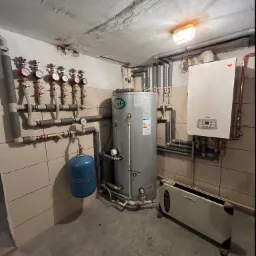 Piwniczne pomieszczenie z zainstalowanym zasobnikiem ciepłej wody Joule, piecem gazowym Termet oraz grzejnikiem kanałowym. Widoczne liczniki ciepła i rury instalacji grzewczej.