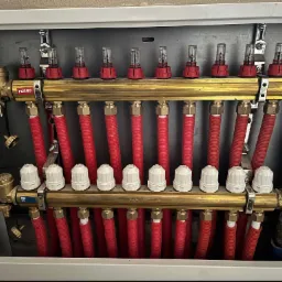 Rozdzielacz ogrzewania podłogowego marki Ferro, z czerwonymi rurami i przepływomierzami, zamontowany w szarej szafce.