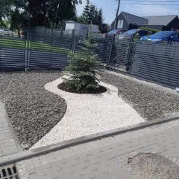 Aranżacja przestrzeni przed domem z ścieżką z białego żwiru prowadzącą do iglaka otoczonego korą, obok szary żwir i metalowe ogrodzenie.