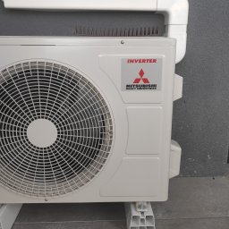 aceco Wojciech Szewczyk - Zewnętrzna jednostka klimatyzacji Mitsubishi Heavy Industries, zamontowana na wspornikach, z kolcami przeciw ptakom na górze, umieszczona na szarej ścianie z oknem.