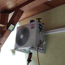 aceco Wojciech Szewczyk - Zewnętrzna jednostka klimatyzacji Mitsubishi Inverter zamontowana na elewacji domu, widoczne rury instalacyjne i narzędzia serwisowe, zadaszenie drewniane z oświetleniem LED w tle.
