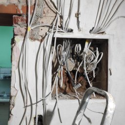 aceco Wojciech Szewczyk - Wewnętrzna instalacja elektryczna w trakcie montażu: wiązka białych kabli wychodzących z otworu w ścianie z odsłoniętą cegłą, fragment drabiny aluminiowej w kadrze.
