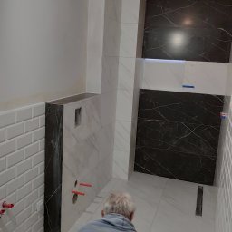 aceco Wojciech Szewczyk - Instalacja płytek w łazience z widocznym pracownikiem, fragmentem ściany z białymi płytkami typu metro, czarnymi płytkami imitującymi marmur oraz odpływem liniowym w podłodze.